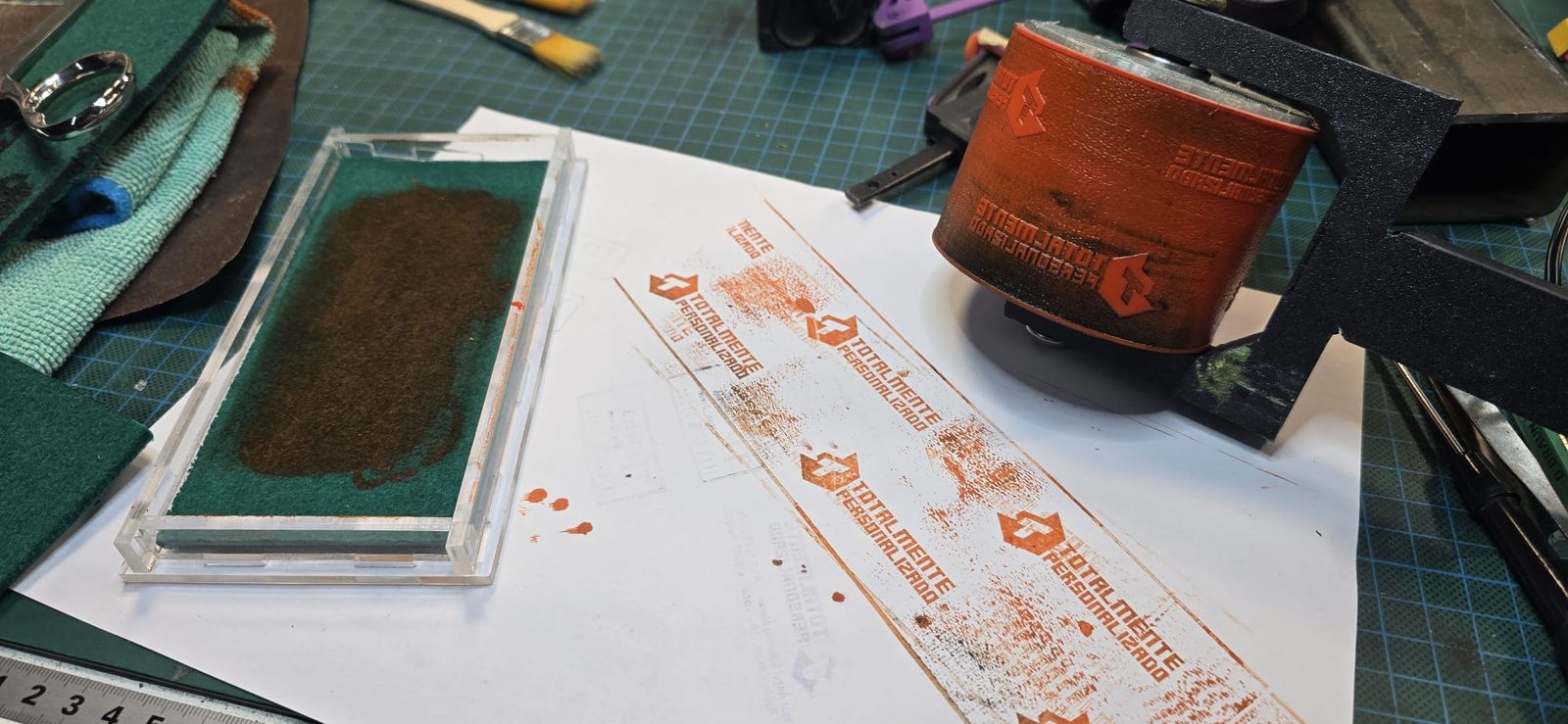 Sello de rodillo montado junto a la bandeja de entintado de metacrilato con fieltro y una tira de papel con los logos estampados en naranja