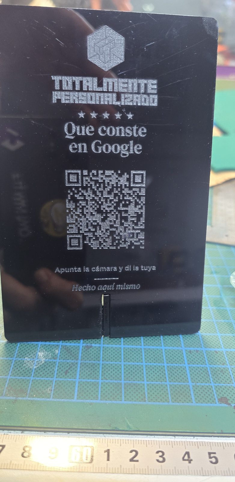 Stand de metacrilato negro de pie sobre una alfombrilla de corte: logo de Totalmente Personalizado, cinco estrellas, el texto Que conste en Google y un código QR grabados en gris claro